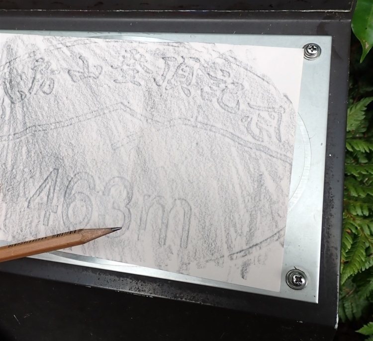 「乳房山登頂記念　463ｍ」と書かれたレリーフの上に白紙を押し当て、上から鉛筆でなぞり石刷りをしている。