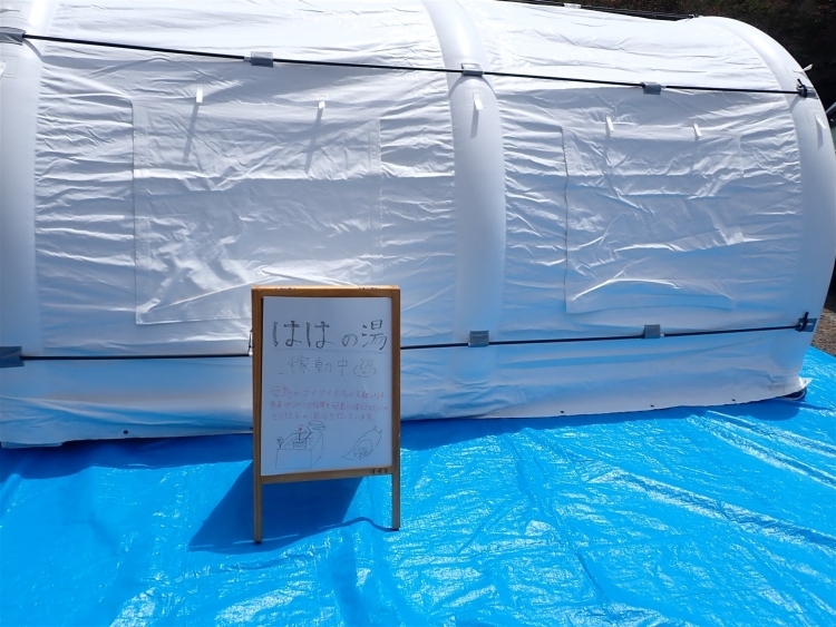 青いビニールシートの上に白いテント。テントの前には「ははの湯」の説明が書かれた看板